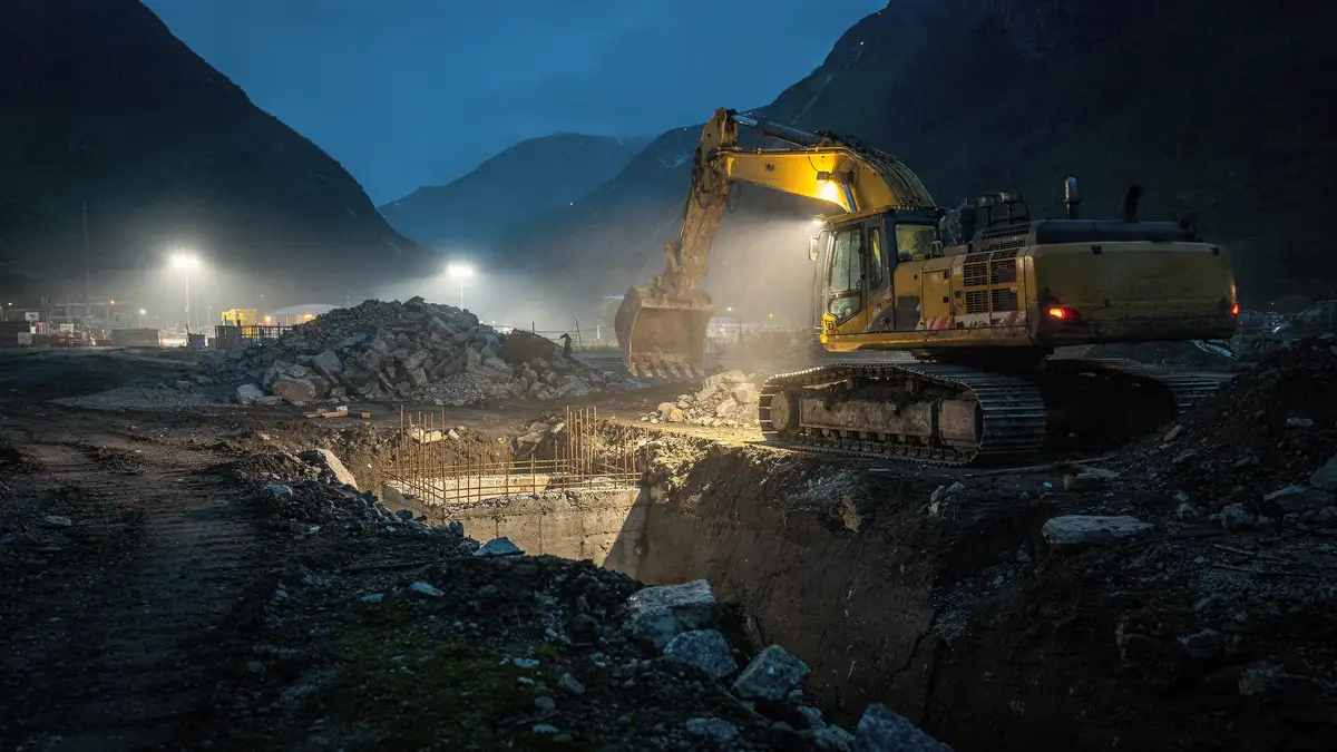 En gravemaskin graver ut en byggegrop på en anleggsplass i skumringen.