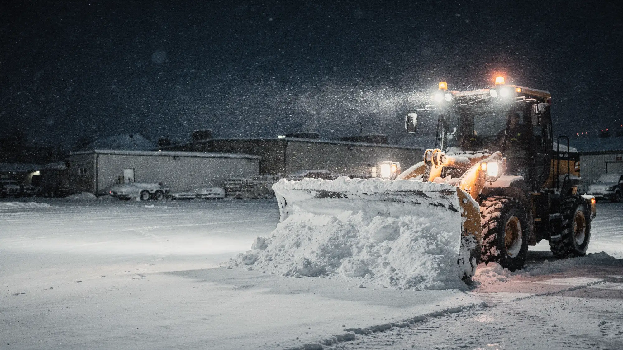 En hjullaster med brøyteskjær rydder snø på en industriplass om natten.